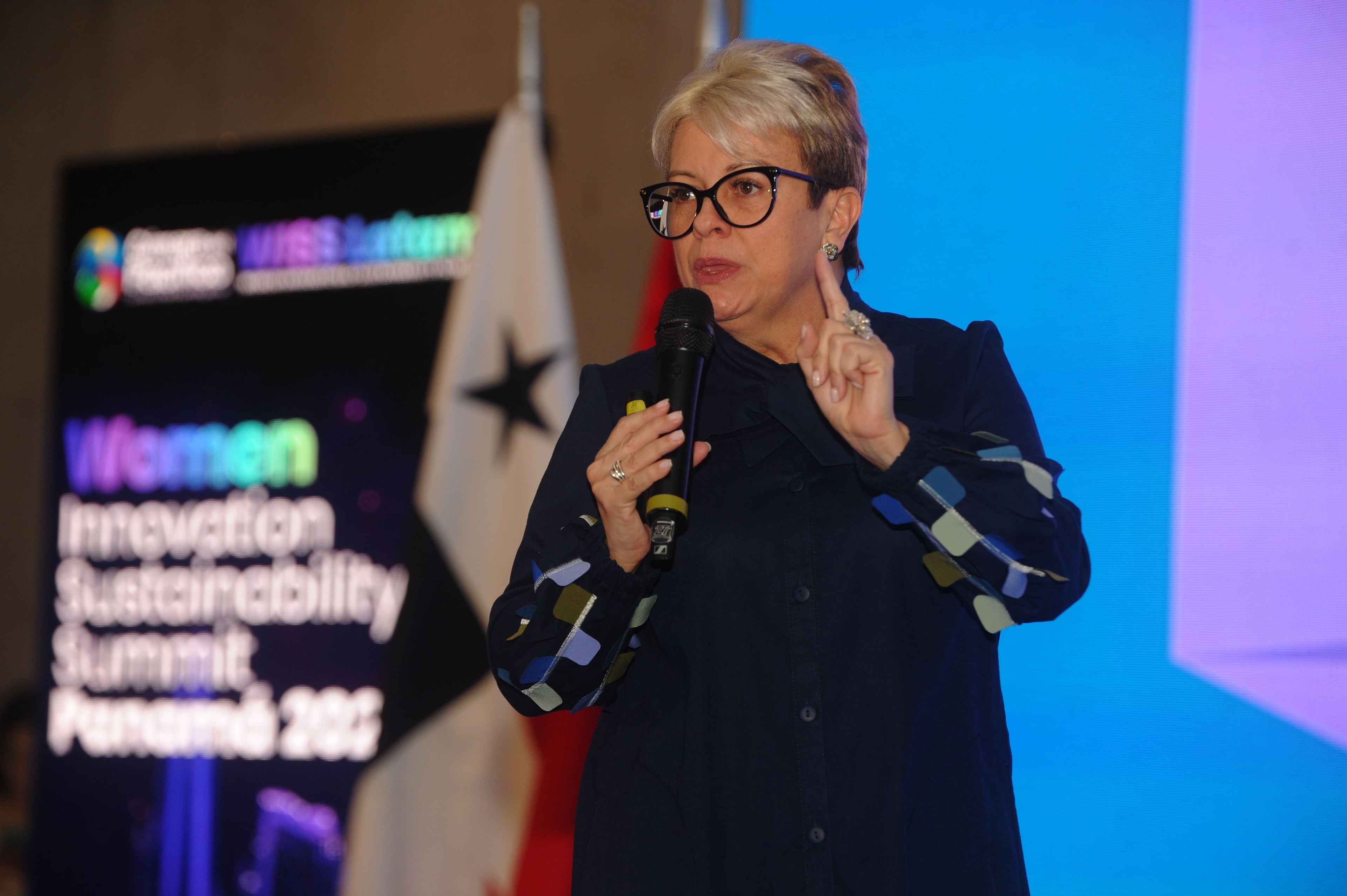 Mujer con lentes hablando con micrófono frente a bandera de Panamá y pantalla WISS Latam — Foro Empresarial 27 Ago 2026