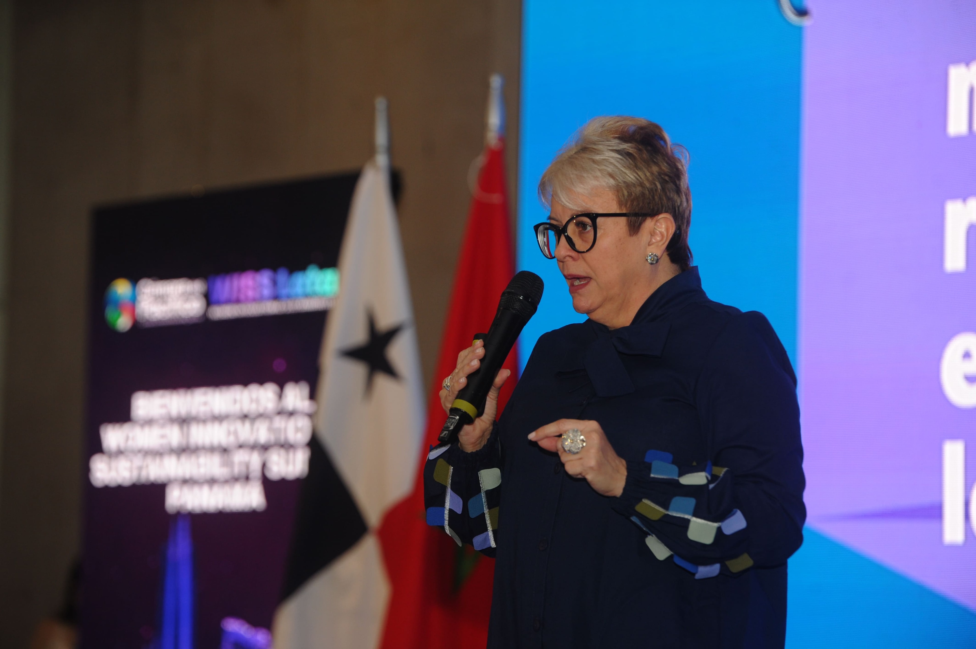 Mujer con lentes hablando con micrófono frente a pantalla "Bienvenidos al WISS" y bandera de Panamá — Foro Empresarial 27 Ago 2026