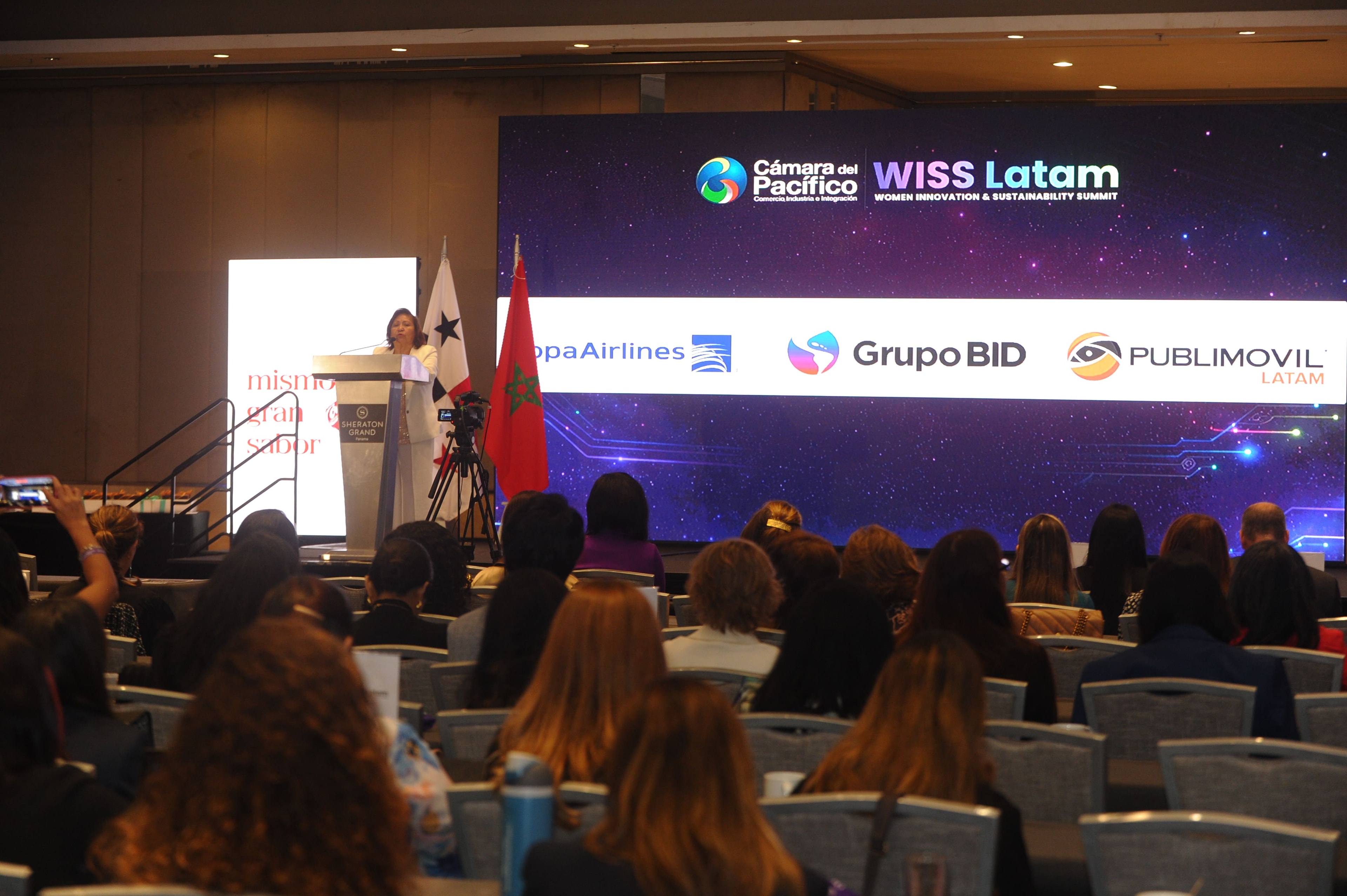 Oradora en el podio con pantalla de patrocinadores Copa Airlines, Grupo BID y Publimovil — WISS Latam 2026